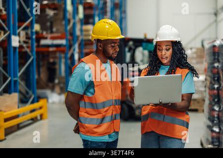 Black female worker wearing hard hat and safety vest showing laptop to male colleague standing in warehouse during work Stock Photo
