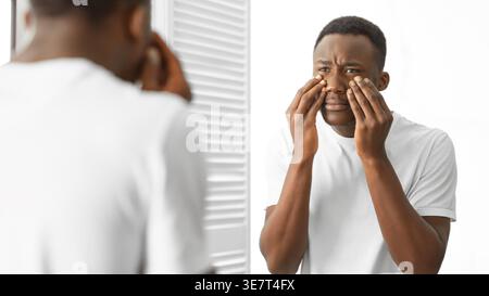 Black man in white shirt looking at the mirror and touching bags under his eyes while standing in the bathroom at home Stock Photo