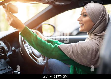 Smiling Middle Eastern woman wearing hijab fixing rear view mirror while driving car during daytime Stock Photo