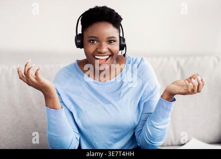 Smiling Black woman with short afro wearing headset spreading hands while sitting on couch in living room at home Stock Photo