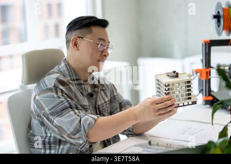 Asian male engineer checking building scale model while sitting at table in office Stock Photo