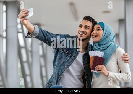 Middle Eastern couple taking selfie using smartphone and holding passport with ticket standing close together in airport Stock Photo
