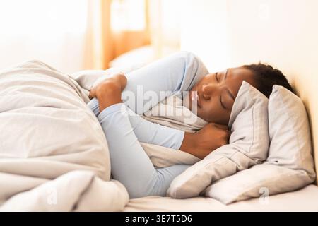 Sleeping Black woman hugging herself with blanket lying in bed at home Stock Photo