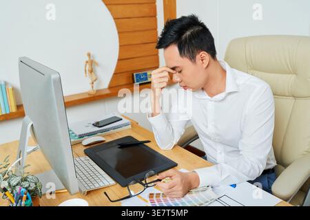 Tired young Vietnamese graphic designer having headache after working on computer for many hours Stock Photo