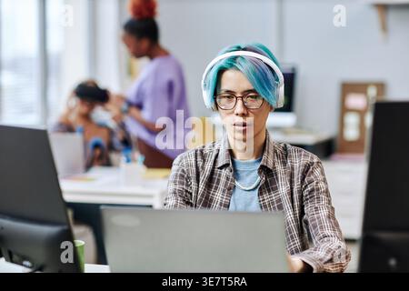 Asian male programmer wearing headset and eyeglasses working on laptop near Black and mixed-race colleagues while sitting at table in office Stock Photo