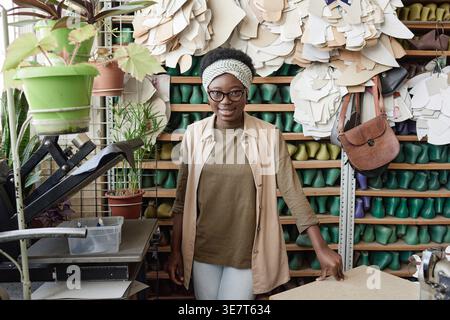 Black female tailor wearing eyeglasses while standing at table in front of shelf in workshop Stock Photo