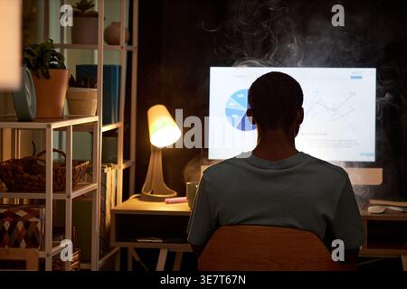 Black male office worker smoking electronic cigarette and working on computer while sitting at desk in office at night Stock Photo