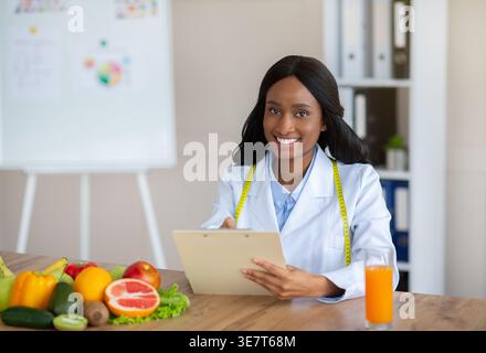 Black female nutritionist with tape measure around neck writing on clipboard sitting at desk in office Stock Photo