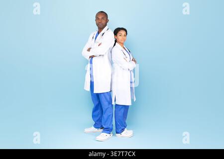 Black male and female doctors in white coat with stethoscope standing back to back with their arms crossed against blue background in studio Stock Photo
