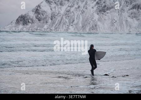 Mixed race man wearing surfing suit holding surfboards while walking on snowy beach during winter Stock Photo