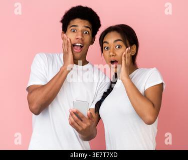 Surprised Black couple touching face with open mouth standing against pink background in studio Stock Photo