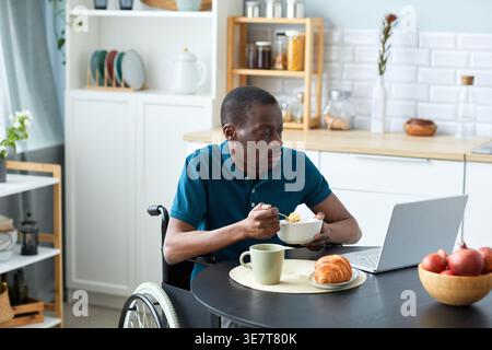 Disabled Black man in wheelchair looking at laptop screen while eating breakfast in kitchen at home Stock Photo