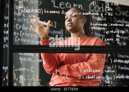 Black female teacher with braids standing near glass board  explaining computer codes in classroom Stock Photo