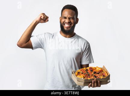 Black bearded man wearing white shirt holding box of pizza and flexing muscles while standing against white background in studio Stock Photo