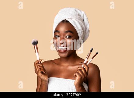 Smiling Black woman wrapped in bath towel holding makeup brushes standing in front of beige background in studio Stock Photo