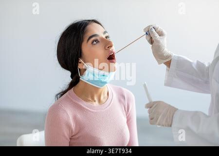 Sick Indian female wearing face mask getting swabbed by doctor while sitting on chair in clinic Stock Photo