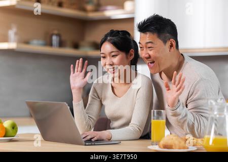 Asian couple having video call waving hands on laptop screen while sitting at table with glasses of orange juice and pastries having breakfast in k... Stock Photo