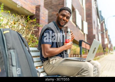 Black male freelancer holding water bottle sitting with laptop on bench on street during daytime Stock Photo