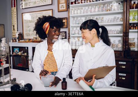 Black female chemist mixing ingredients using mortar and pestle standing beside Japanese female colleague holding chart in vintage pharmacy Stock Photo