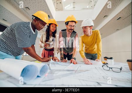 Busy Diverse group of architects wearing safety helmets working on blueprint on the table in the office Stock Photo