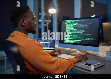 Black male programmer typing codes on computer while sitting at table with smartphone working late at night in office Stock Photo
