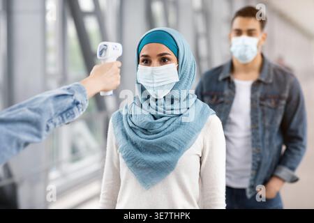 Hand of airport worker checking temperature of Middle Eastern woman using electronic thermometer near boyfriend standing in airport Stock Photo
