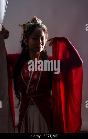Asian woman in traditional red dress wearing flower crown pulling curtain aside while standing with raised hand near face against purple taupe back... Stock Photo