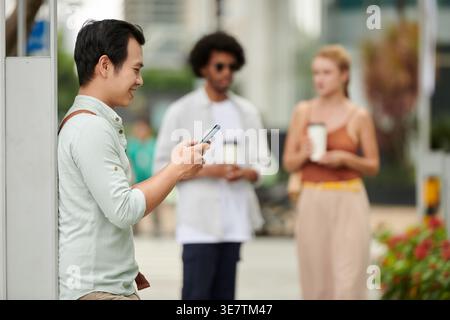 Asian man using smartphone standing and leaning on post outdoors during daytime Stock Photo