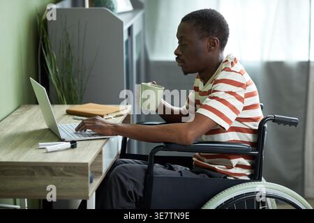 Disabled Black man in wheelchair using laptop and drinking coffee working from home Stock Photo