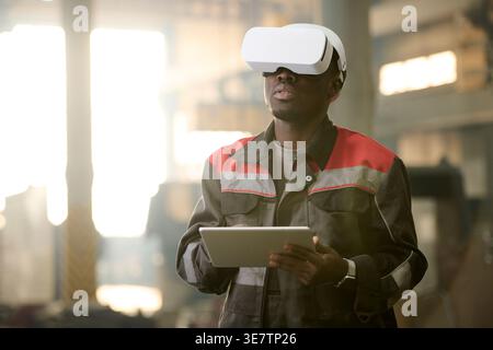 Black male factory worker in uniform wearing VR glasses using tablet while standing in workshop Stock Photo