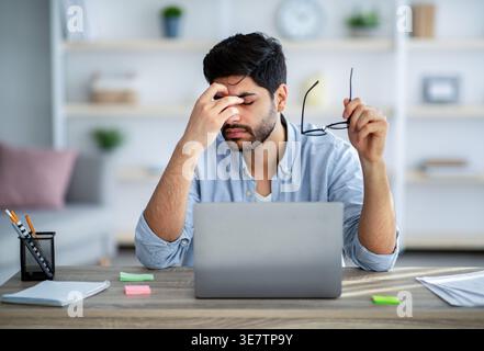 Tired Arab man with beard holding eyeglasses sitting at table with laptop at home Stock Photo