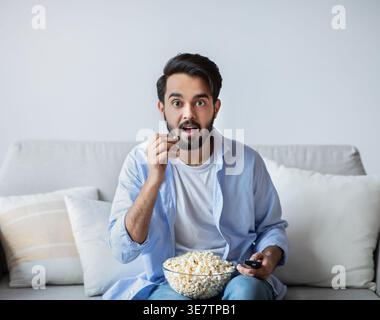 Middle Eastern man eating popcorn sitting on sofa in living room at home Stock Photo