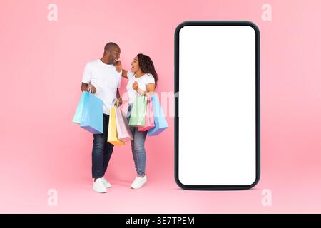 Cheerful Black couple with colorful shopping bags looking at each other while standing near huge smartphone against pink background in studio Stock Photo
