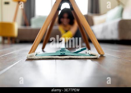 Hispanic female teenager wearing rubber gloves using mop in cleaning wooden floor under the chair in living room at home Stock Photo