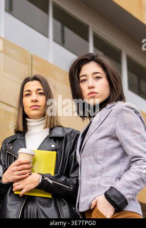 Hispanic business woman standing with caucasian female colleague holding coffee and yellow envelope outside the office building during break time Stock Photo