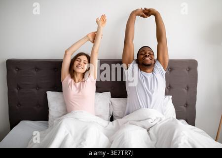 Black and caucasian couple waking up stretching their arms while sitting on bed at home Stock Photo