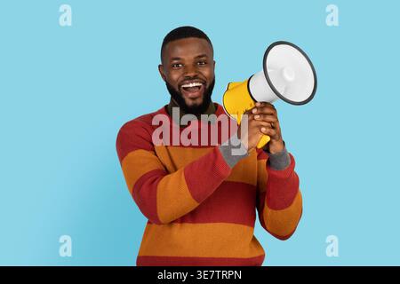 Smiling Black man holding megaphone while standing against blue background in studio Stock Photo