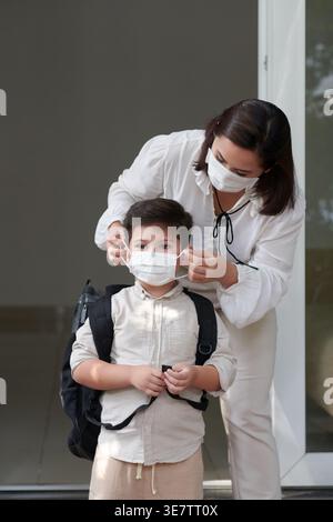 Asian mother helping Mixed race son wearing medical mask getting ready for school standing outside house during daytime Stock Photo