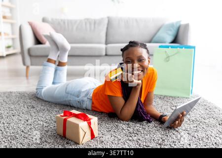 Cheerful Black woman paying online using credit card on tablet while lying on carpet with shopping bags and parcel box in living room at home Stock Photo