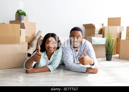 Smiling Black couple showing thumbs up while lying on floor among cardboard boxes in living room at home Stock Photo