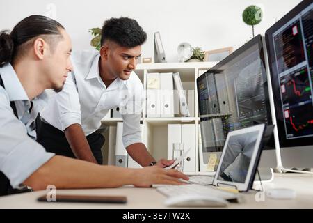 Indian male trader talking to mixed-race male colleague sitting at table in office Stock Photo