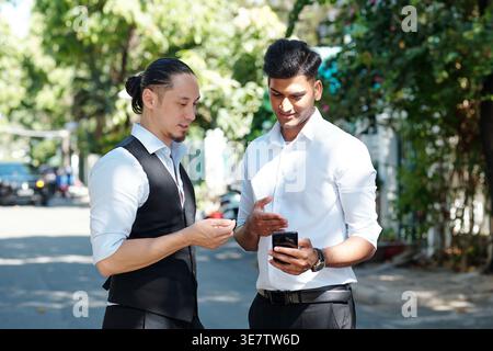 Indian man showing smartphone to his mixed-race male friend while standing outdoors during daytime Stock Photo