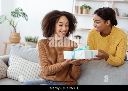 Black woman giving gift box to female friend while sitting on sofa in living room at home Stock Photo