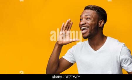Attention. Funny African American Man Shouting Holding Hand Near Mouth On Yellow Studio Background. Panorama, Copy Space Stock Photo