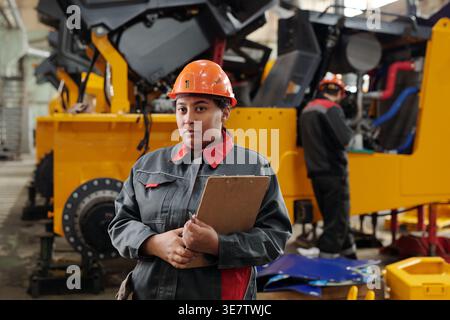 Serious Mixed race female engineer wearing safety helmet holding clipboard standing in front of caucasian male coworker in factory Stock Photo