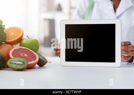 Cropped image of Black female nutritionist holding tablet with blank screen sitting at table with fruits and vegetables in clinic Stock Photo
