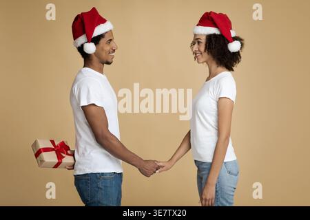 Black man wearing Santa hat holding giving gift box with red ribbon behind his back while standing facing girlfriend holding hands against brown ba... Stock Photo