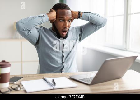 Shocked Black businessman touching his head and looking at laptop screen while sitting at table with smartphone, eyeglasses, disposable coffee cup,... Stock Photo