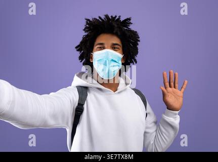 Black man with afro wearing white hoodie and face mask taking a video and waving his hands while standing against purple wall in studio during pand... Stock Photo