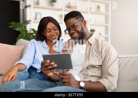 Black woman looking at husband using tablet sitting close together on sofa at home Stock Photo